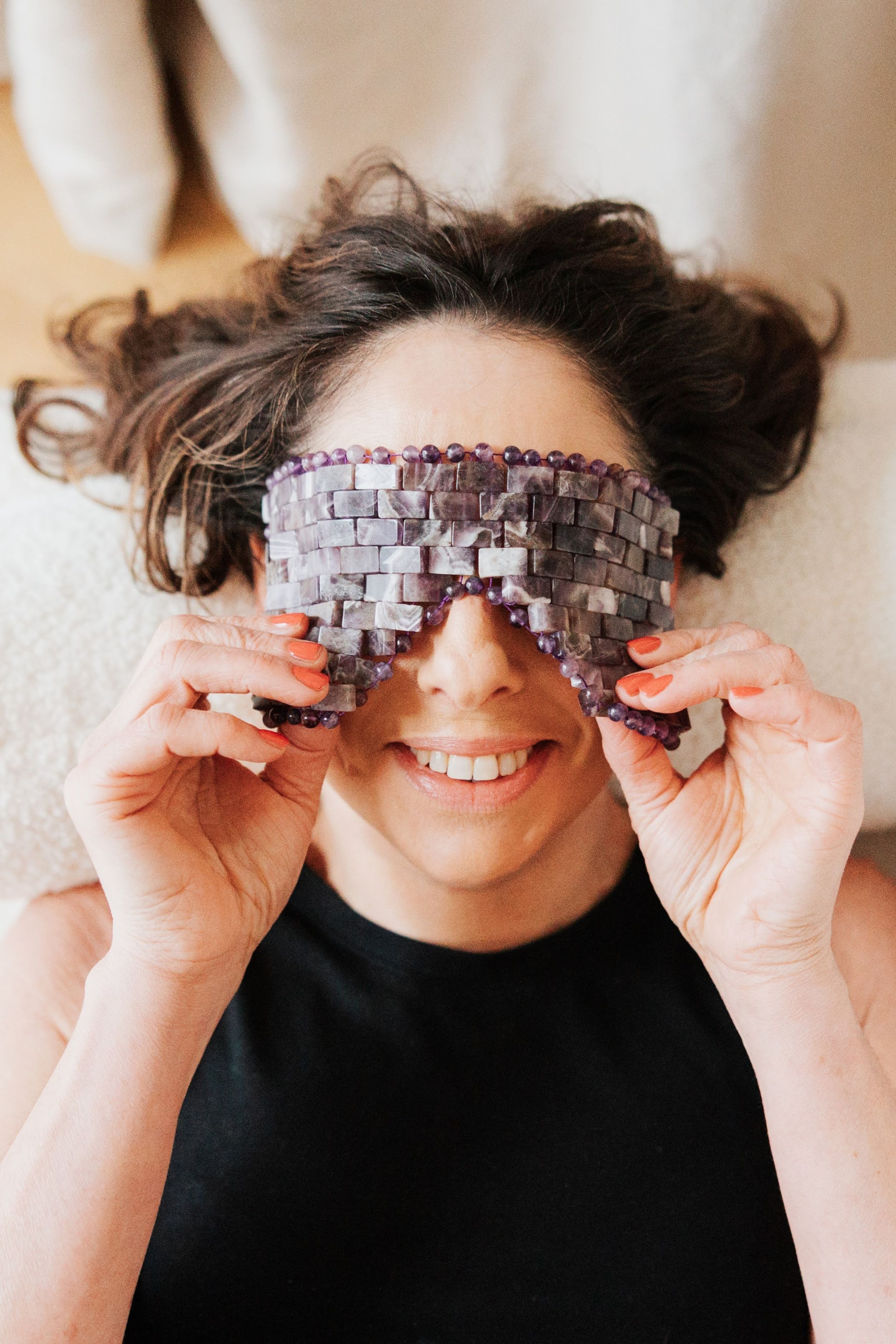 Smiling woman lying down holding a crystal amethyst eye mask over her eyes during a spa treatment.