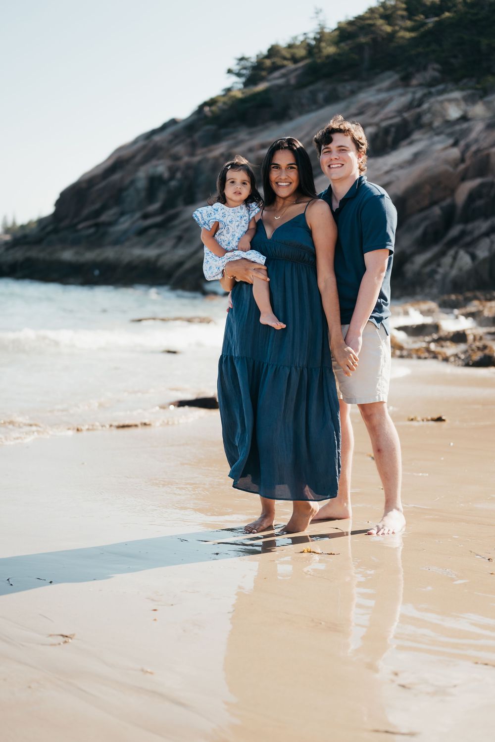 A family stands barefoot at Sand Beach Acadia with their toddler.
