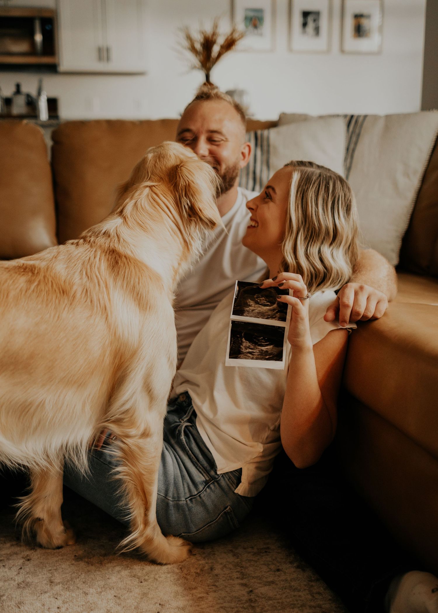 A couple sitting on floor with golden retriever sharing ultrasound photo in warm-toned living room setting.