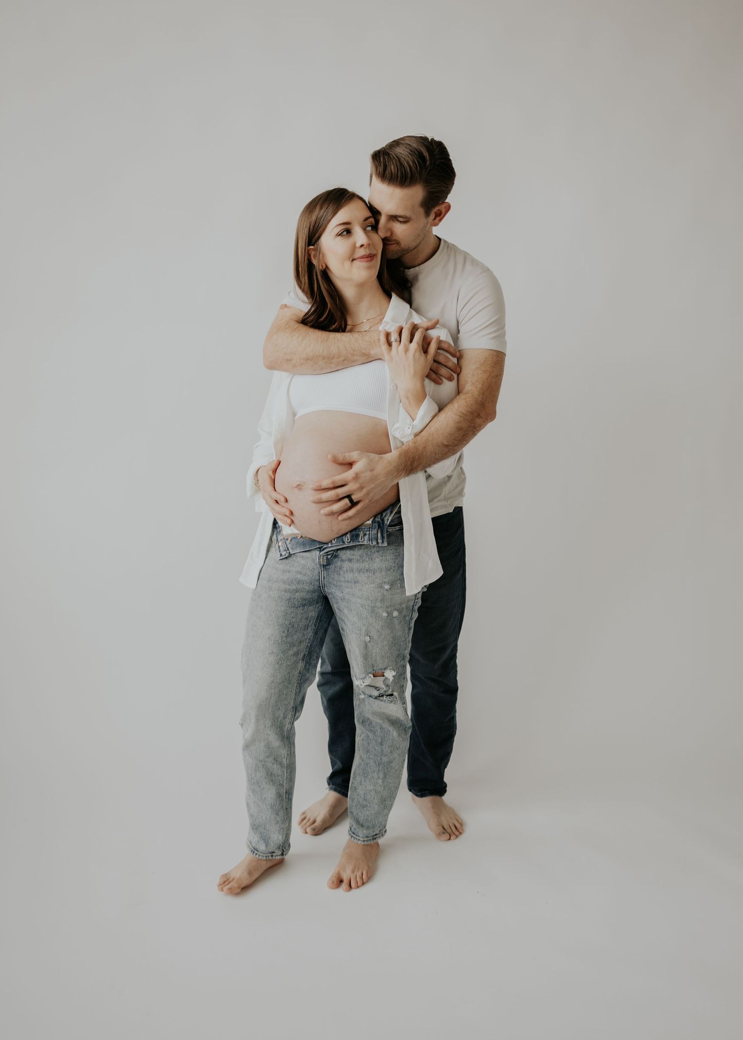 Minimalist maternity photo featuring couple in casual clothing embracing against white background.