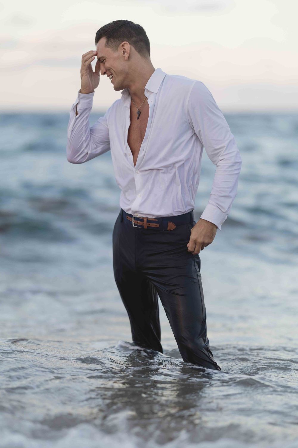 Person in white shirt and dark pants standing in ocean waves at sunset.