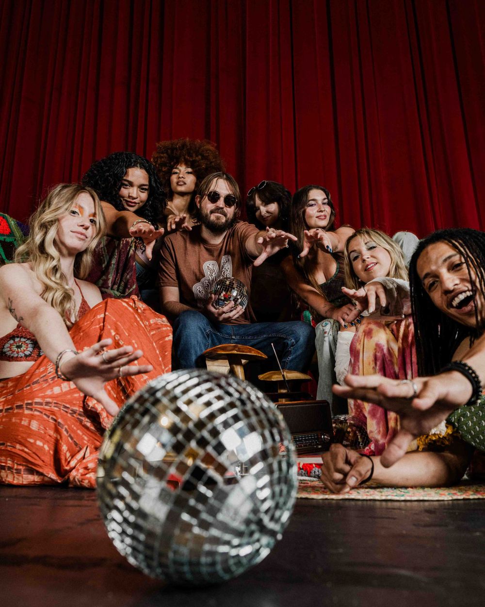 Group poses casually with a disco ball in front of red curtains at a theater venue.