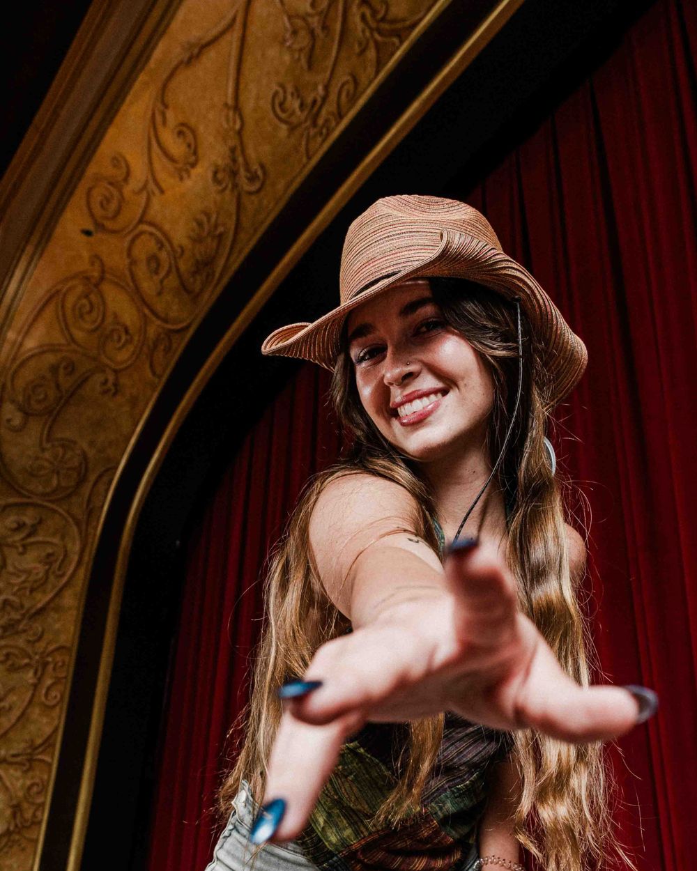 Person in a pink hat reaches towards camera in front of ornate theater architecture.