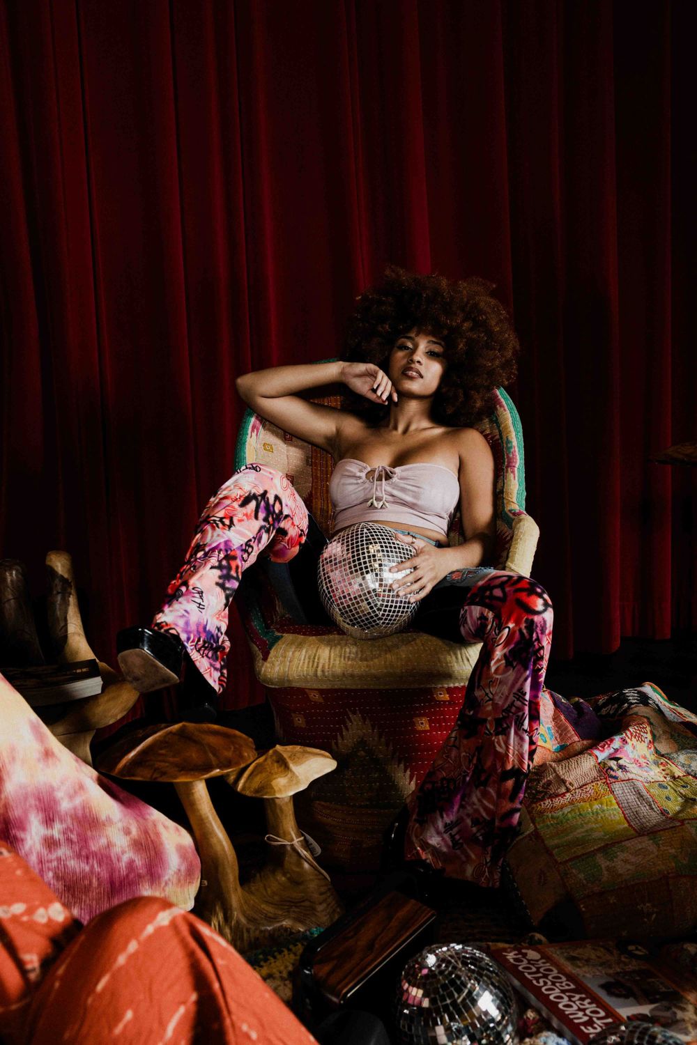 Person lounges in vintage chair wearing sparkly dress and colorful boots against red curtain.