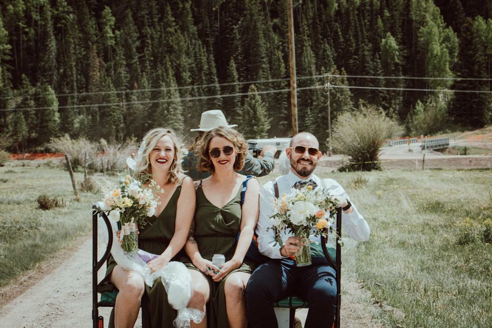 Three people sitting on a bench outdoors with flowers and evergreen trees in the background.