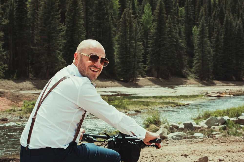 Person in suspenders sits by a mountain stream while wearing sunglasses.