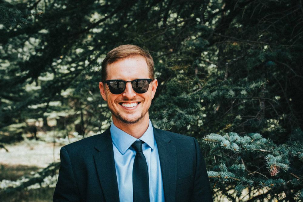 Outdoor portrait of someone in a navy suit and sunglasses with trees in background.