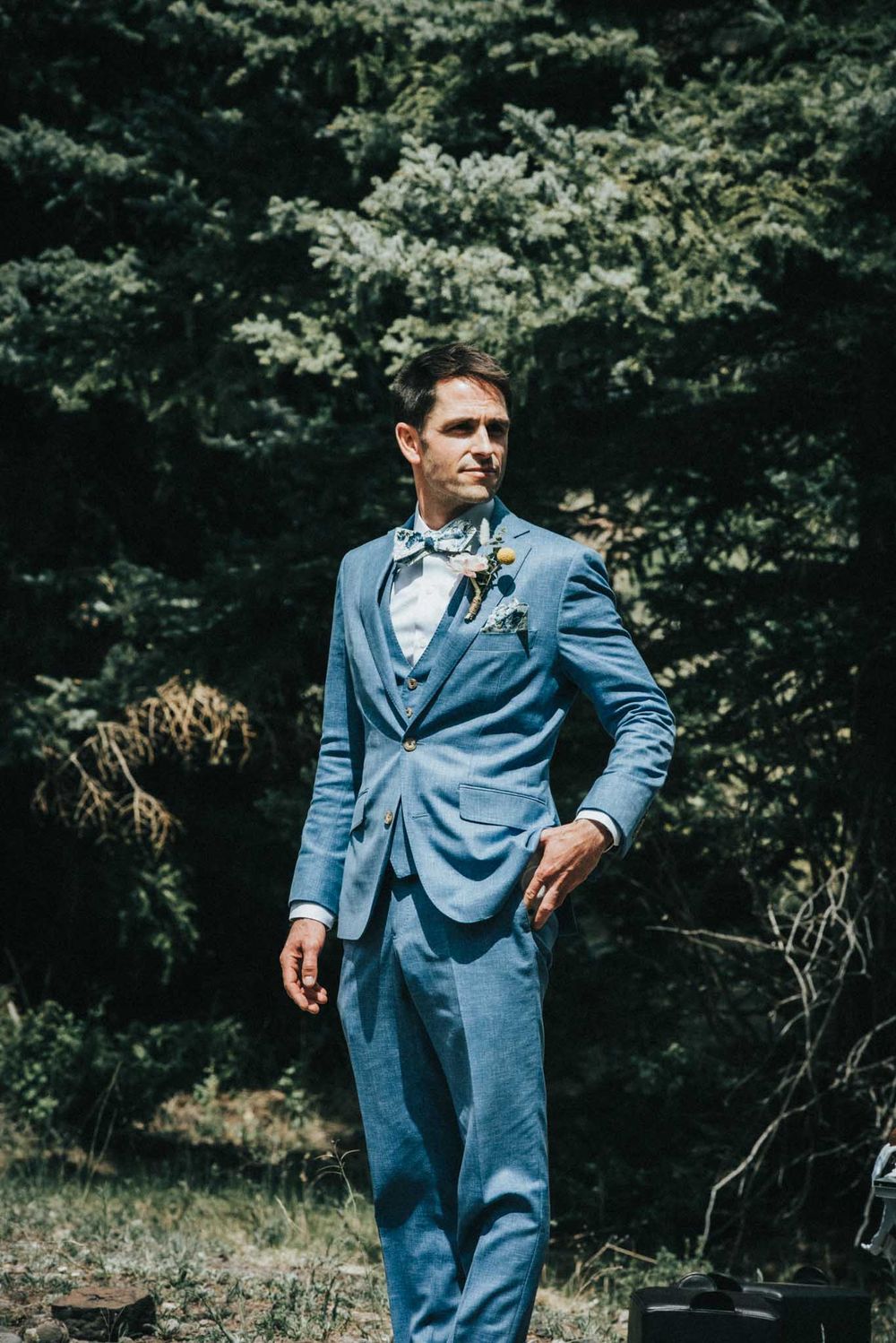 Groom in a light blue suit with bow tie poses in front of forest backdrop.