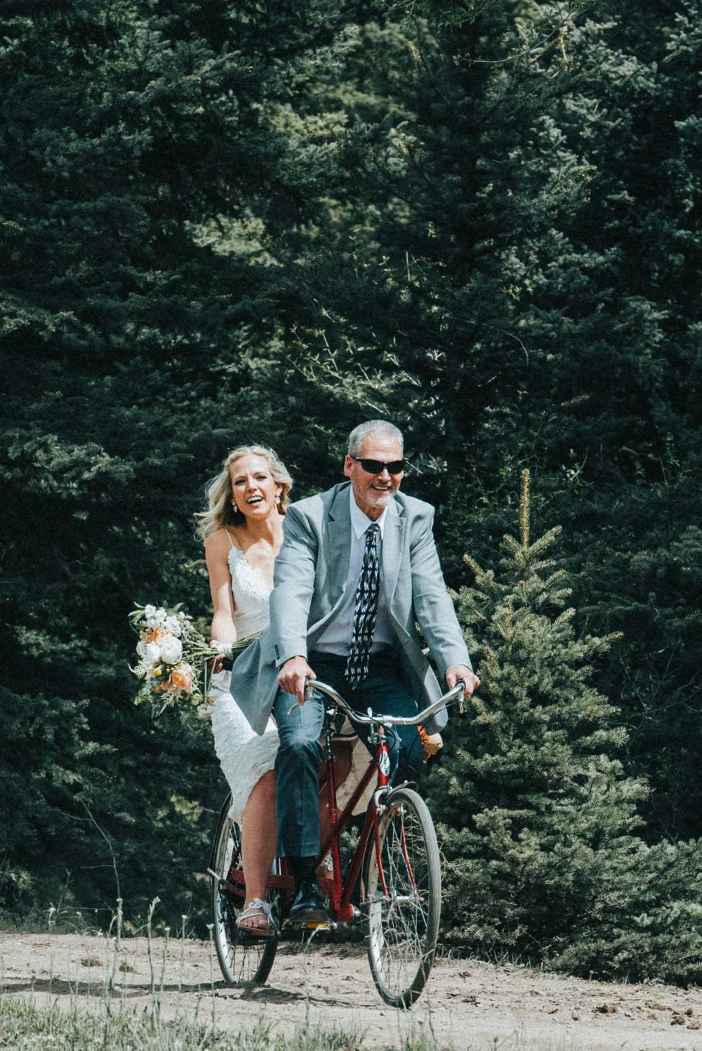 Wedding couple enjoying a playful bicycle ride against a backdrop of evergreen trees.
