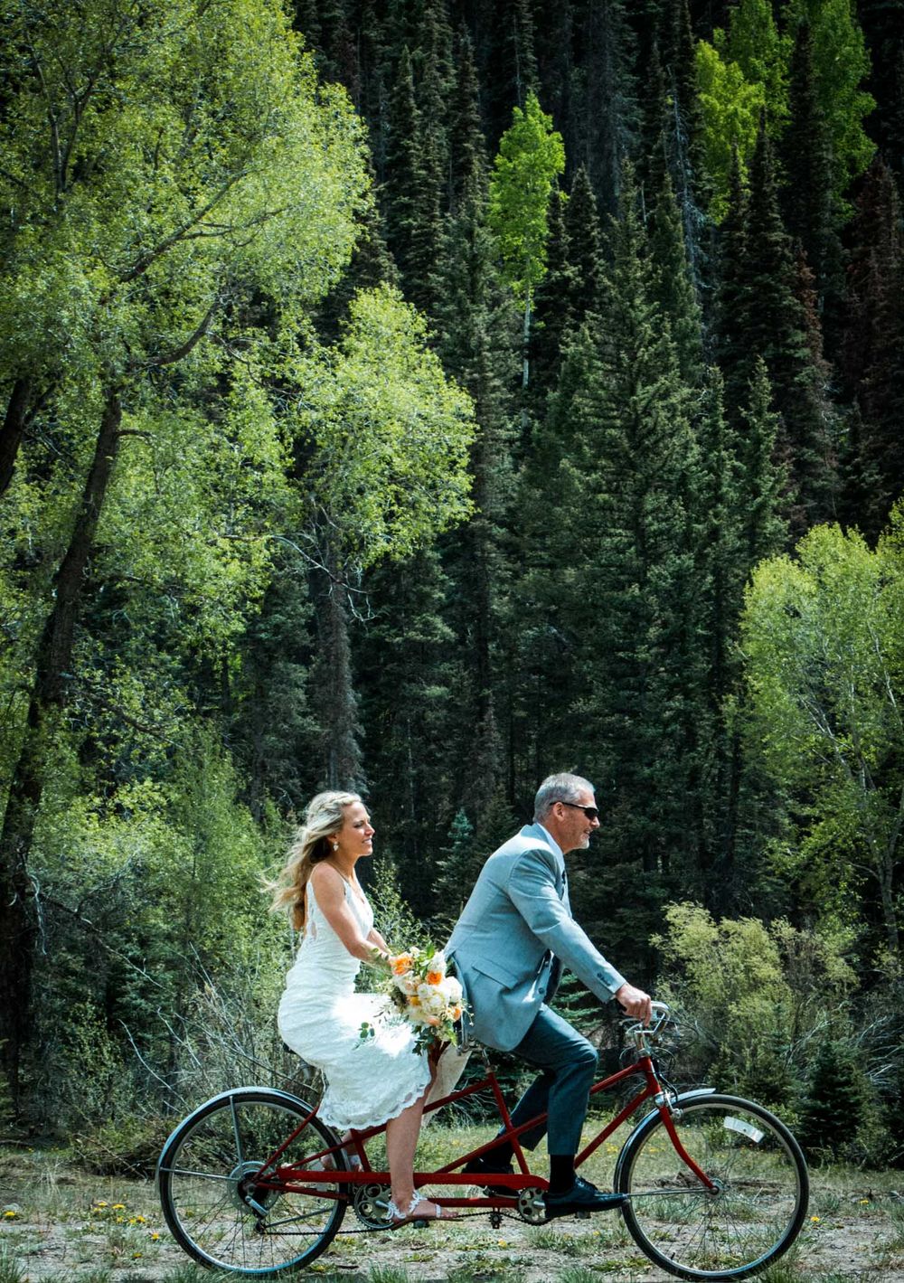 A tandem bicycle ride through a lush green forest setting during a wedding celebration.