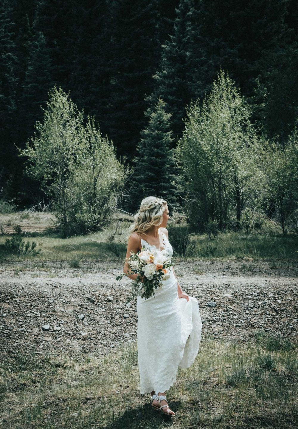 A figure in a flowing white wedding dress stands among evergreen trees holding a floral bouquet.
