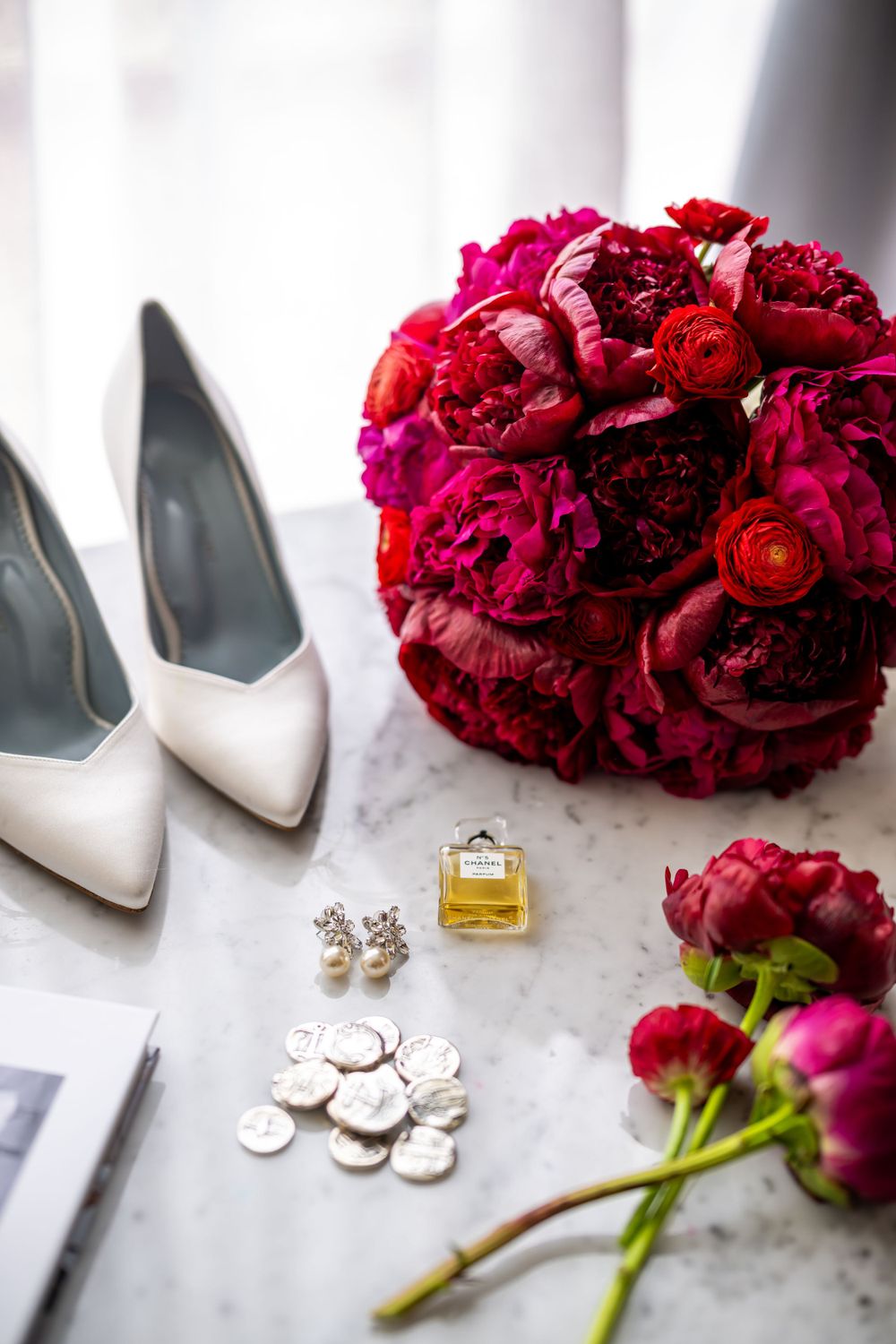 Wedding detail flatlay with white heels, deep red rose bouquet, perfume and jewelry on marble surface.