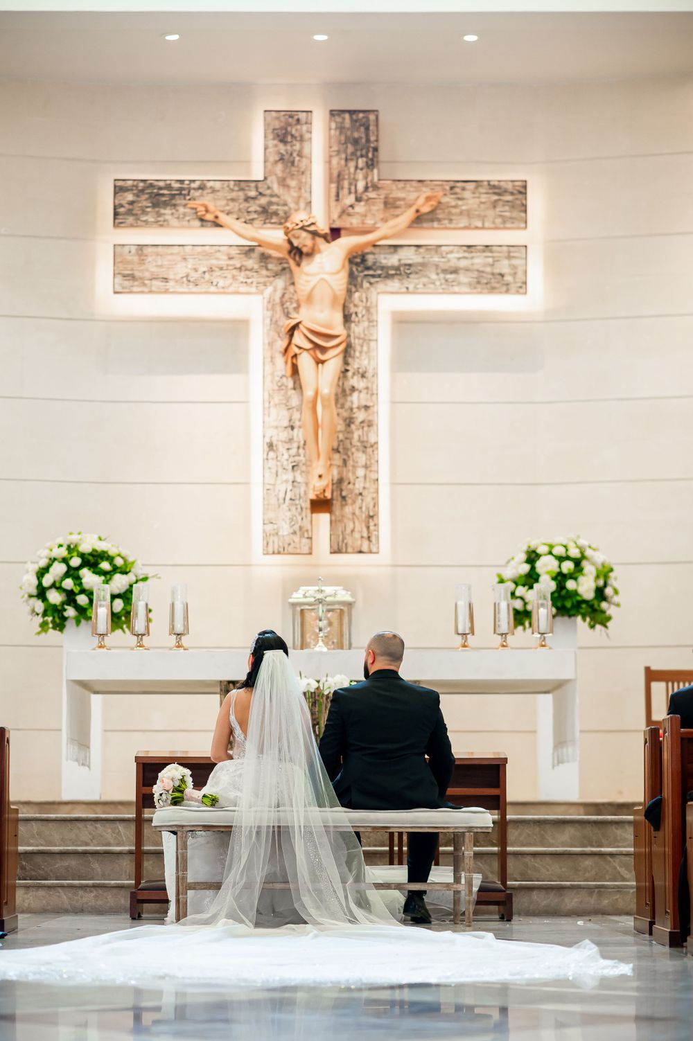 Wedding couple seated at altar beneath large crucifix in white church.