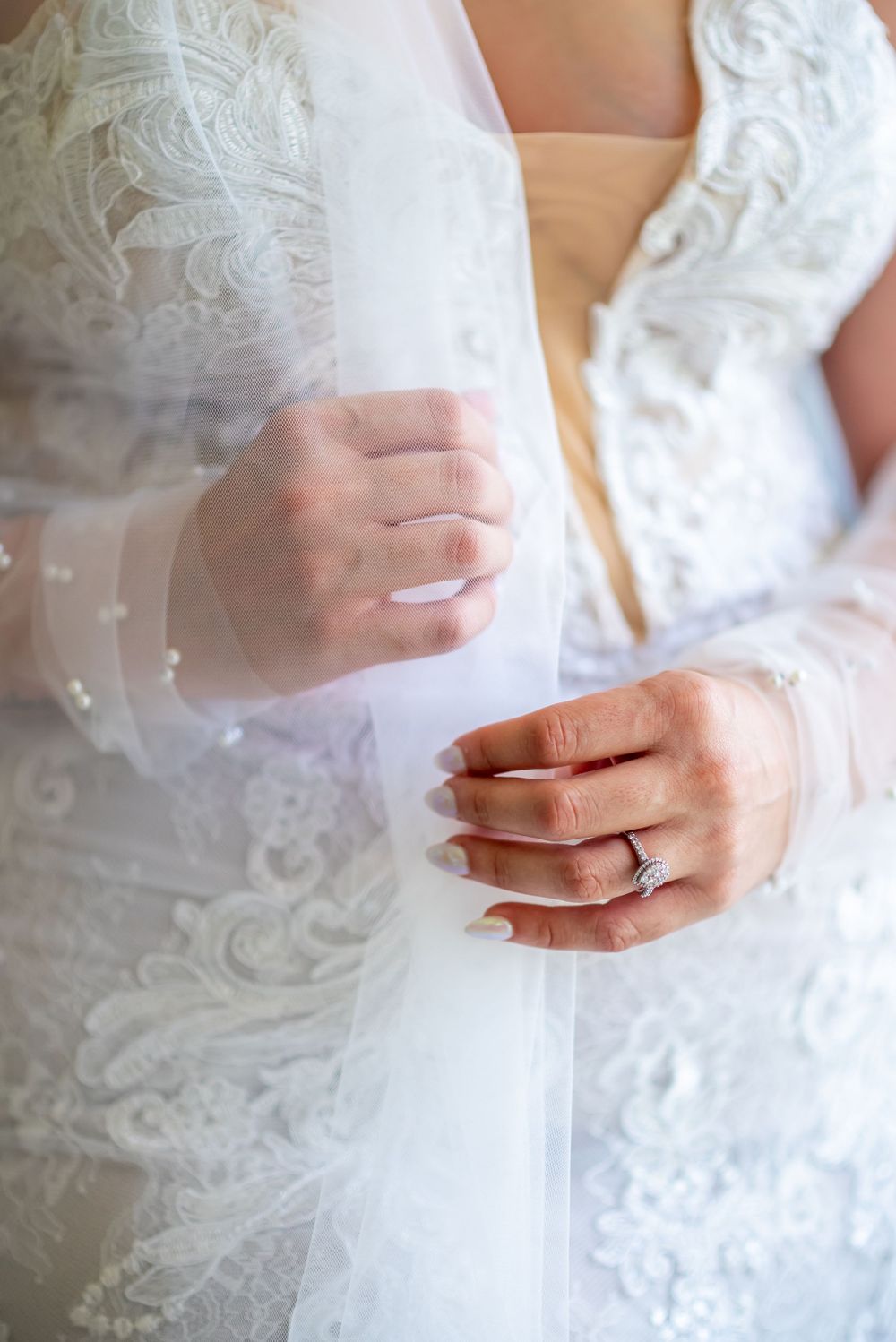 Close-up of bride's hands holding delicate veil, showing wedding ring and lace dress details.