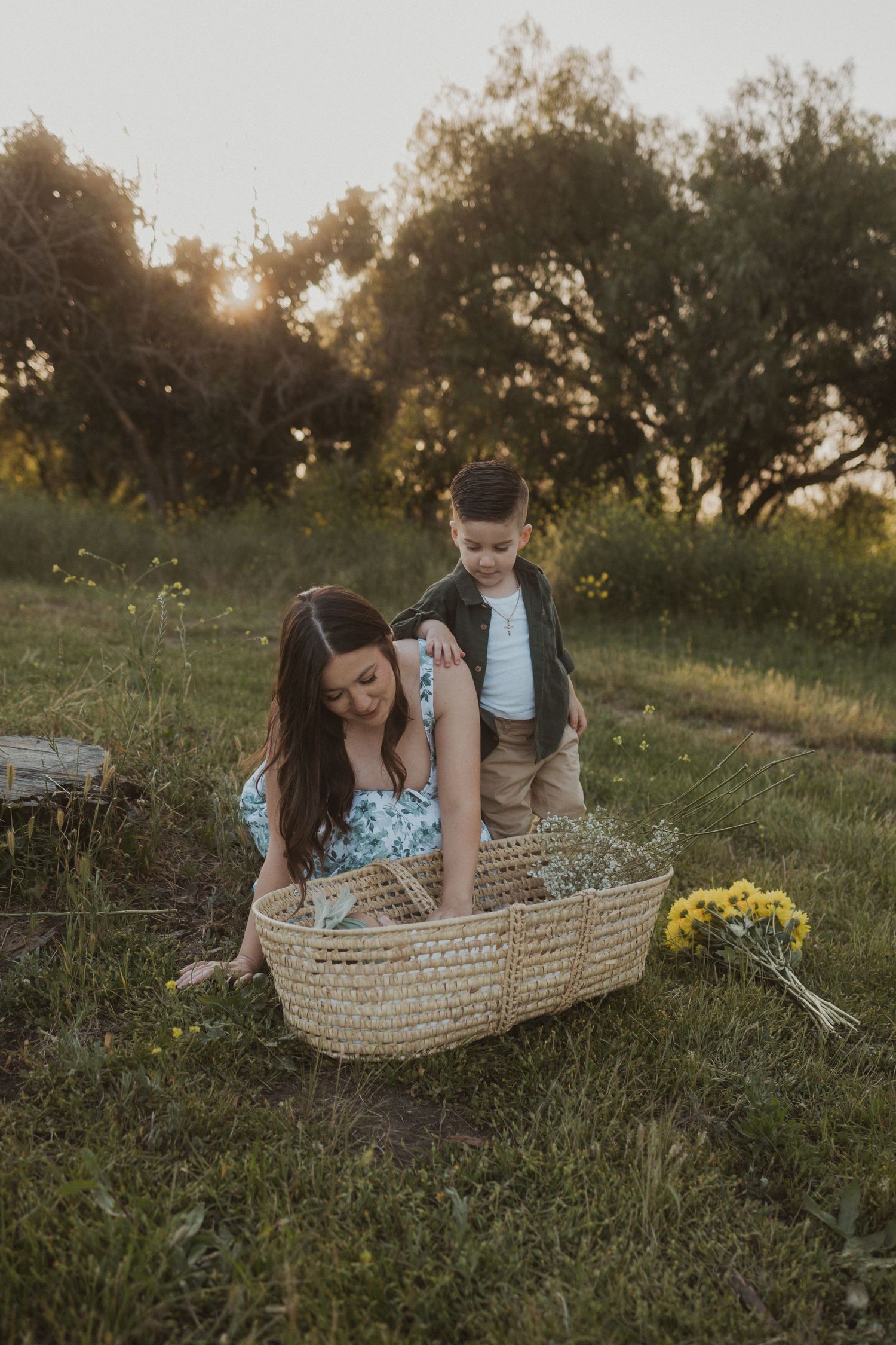 Woman kneeling on grass gently touching her baby in a basket with a young boy standing beside her.
