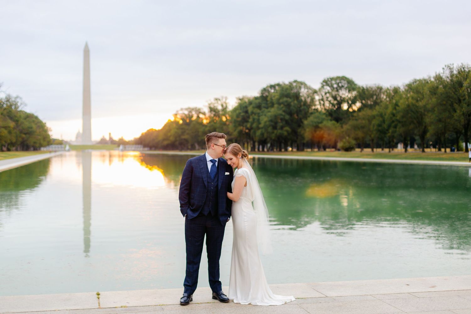 Bride and groom standing at reflecting pool with Washington Monument at golden sunrise in background.