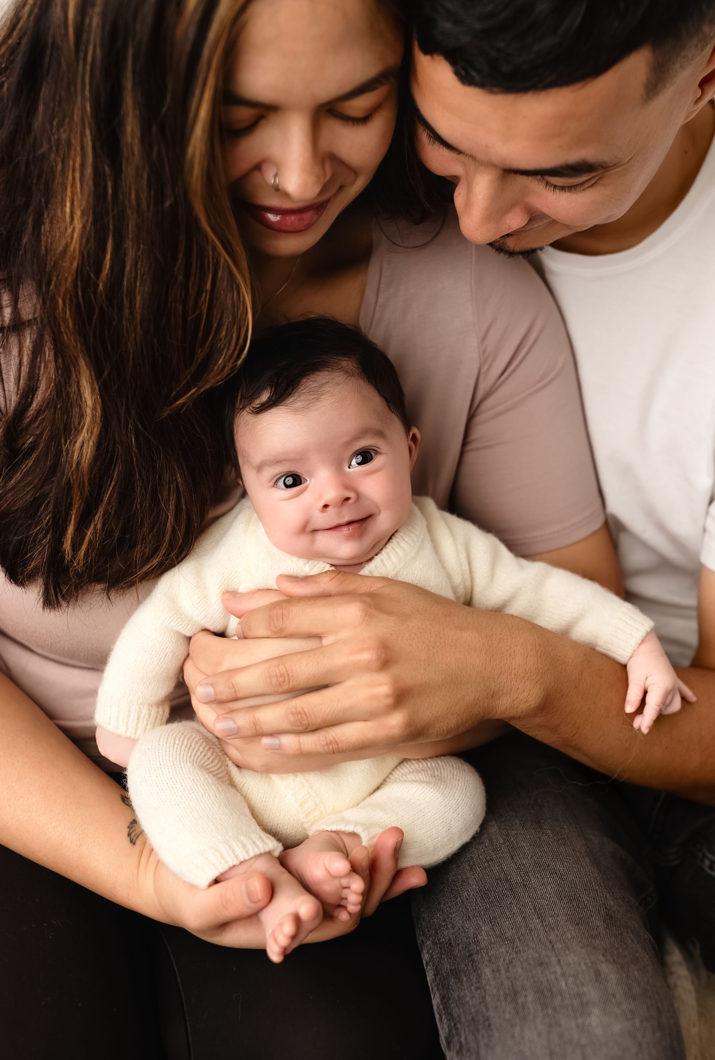New parents snuggle their smiling newborn girl, who is dressed in a knit cream, long-sleeved romper, by Charlottesville baby photographer.