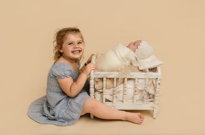 Adorable big sister with blond curls holds her baby's spindle crib and smiles proudly, captured by Charlottesville newborn photographer.