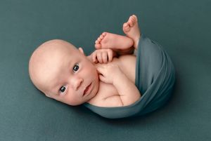 Bright-eyed baby boy lies, partially wrapped, against a turquoise blanket during his newborn session by Charlottesville family photographer.