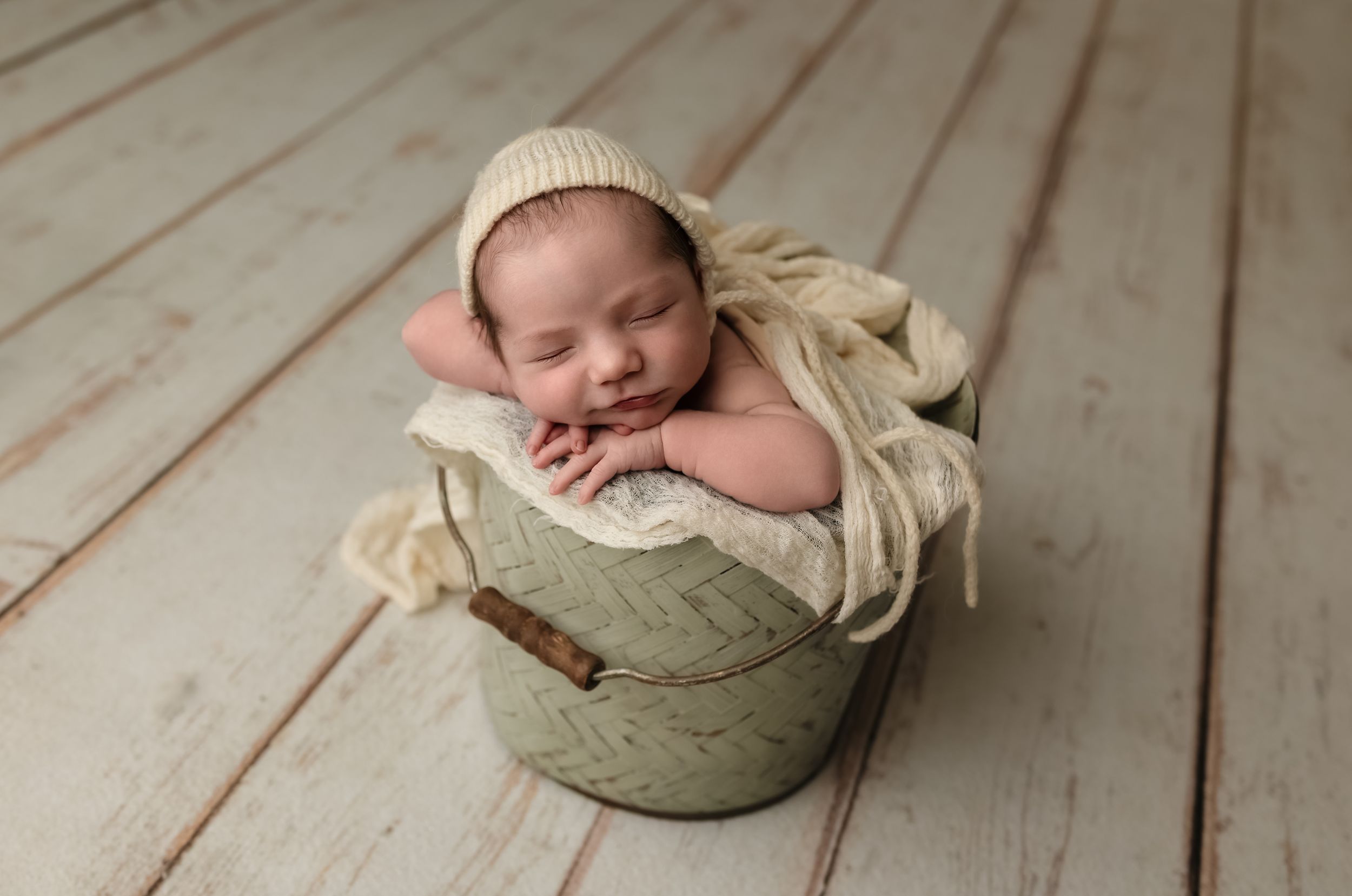 Newborn smiles in a bucket against a white, wood background and dressed in a cream knit hat, by Charlottesville baby photographer.