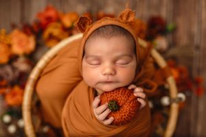 Baby girl sleeps holding a pumpkin in a bear hat and orange wrap against a fall floral backdrop by Charlottesville newborn photographer.