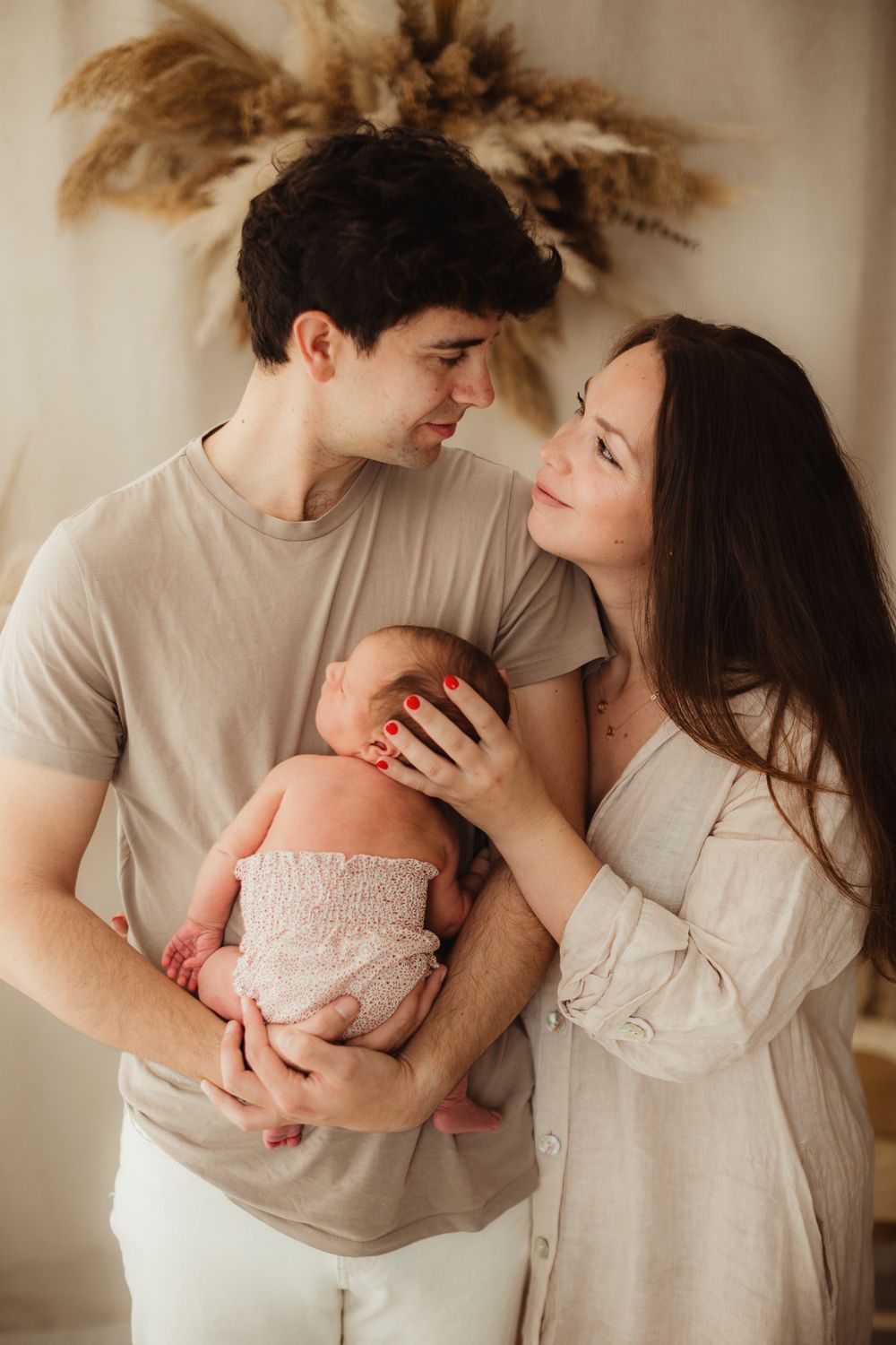 Young couple in neutral tones lovingly holding their newborn baby in a warm, softly lit boho-style room.