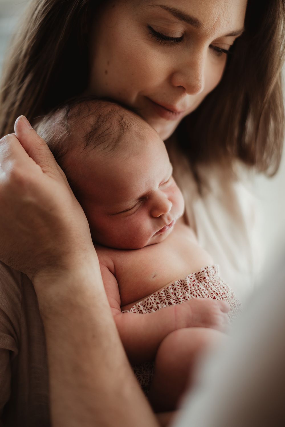 A tender mother gently cradles her sleeping newborn baby close to her chest in a soft, intimate portrait.