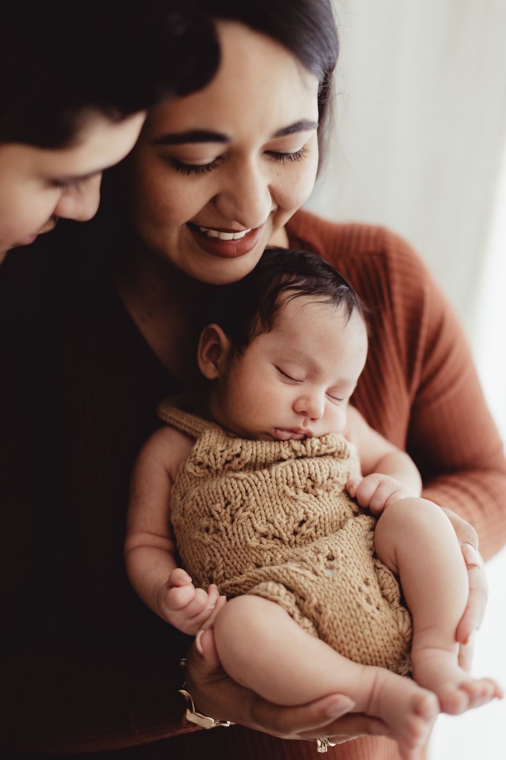 Parent gently cradles newborn baby wearing soft knit sweater in intimate indoor photoshoot series.