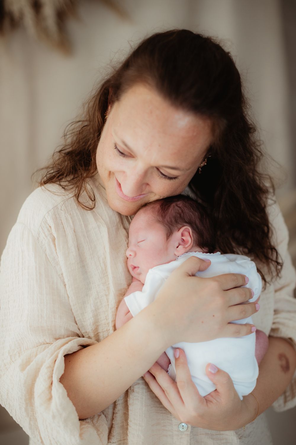 A smiling mother with curly dark hair tenderly holds her sleeping newborn baby dressed in white.