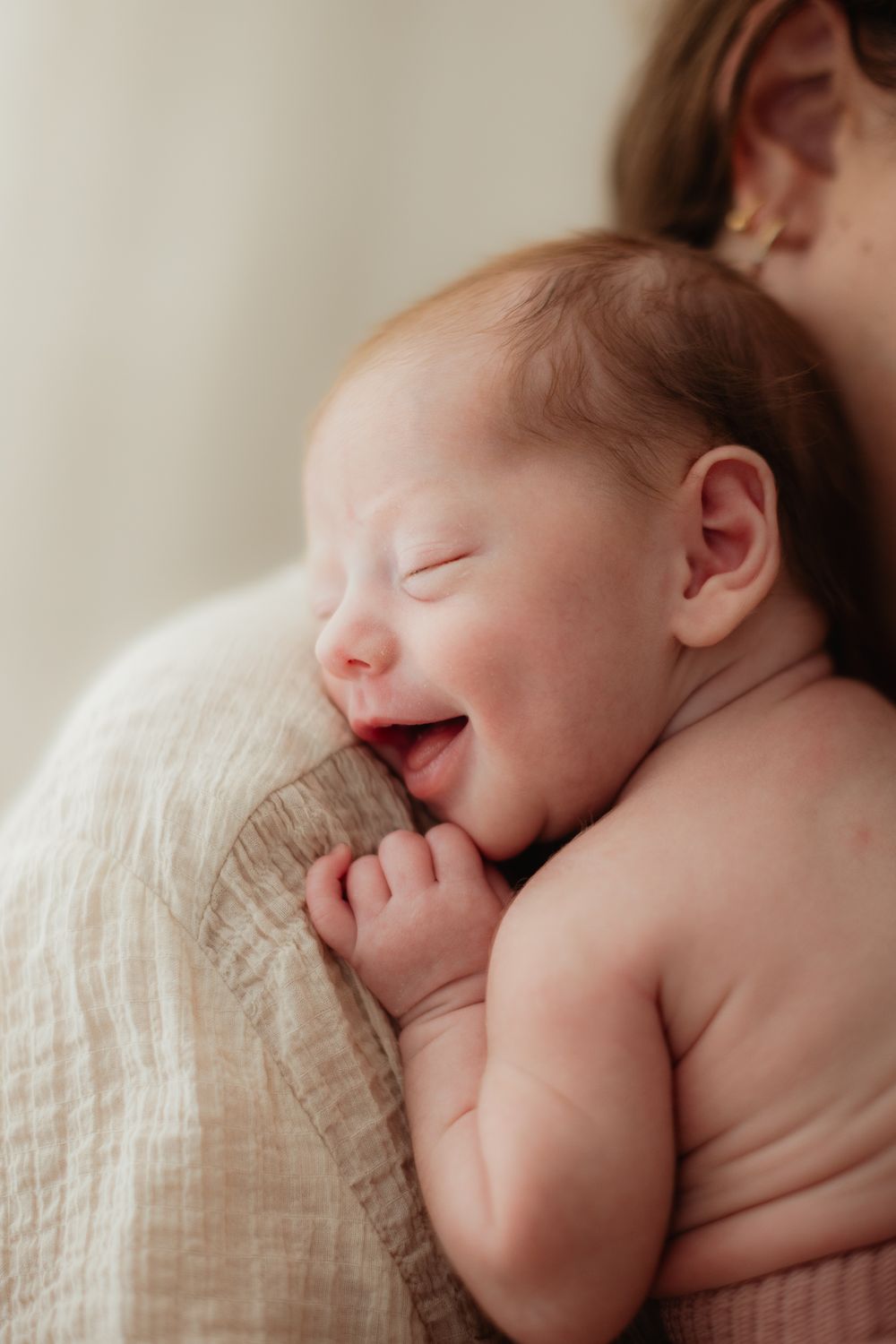 Newborn baby smiling while being held against mother's shoulder, wrapped in a soft muslin cloth.