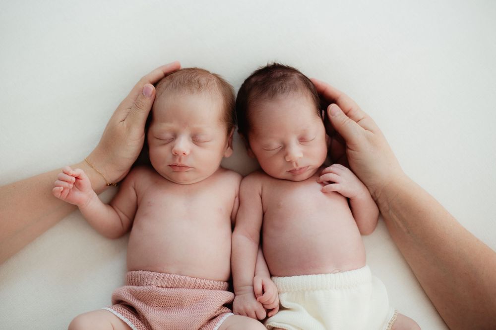 Two sleeping newborn twins lying side by side, gently cradled by parents hands on a white background.