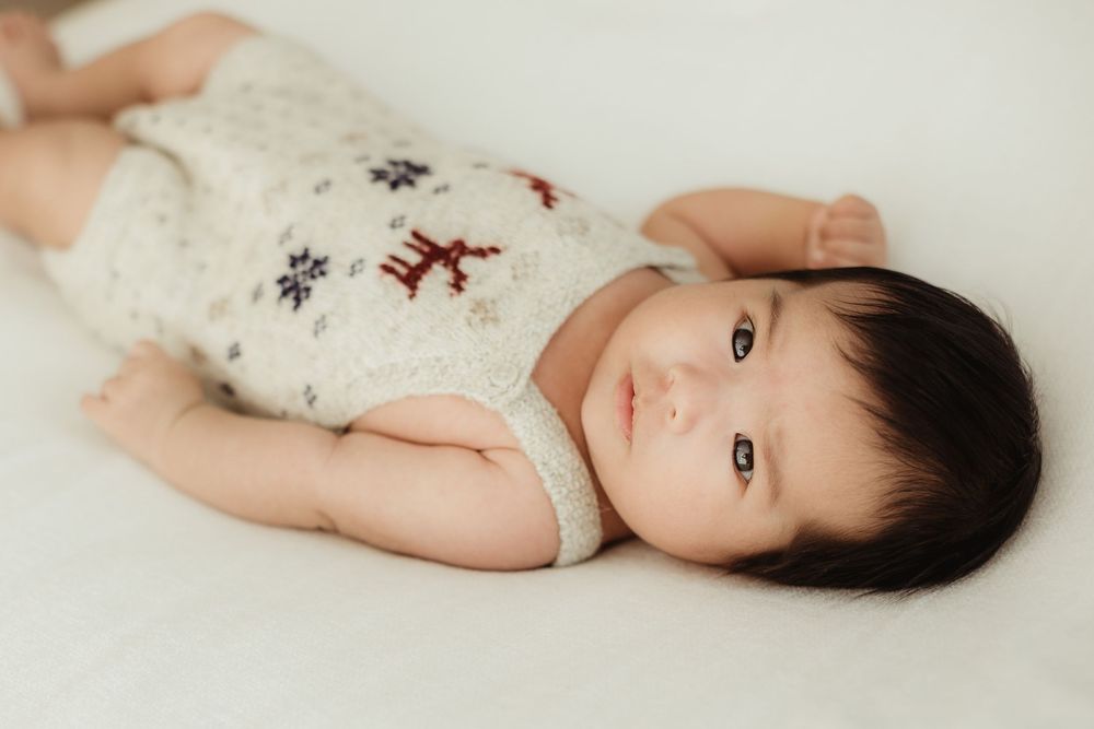 A baby in a white floral outfit lies on a soft cream-colored surface in a series of gentle poses.