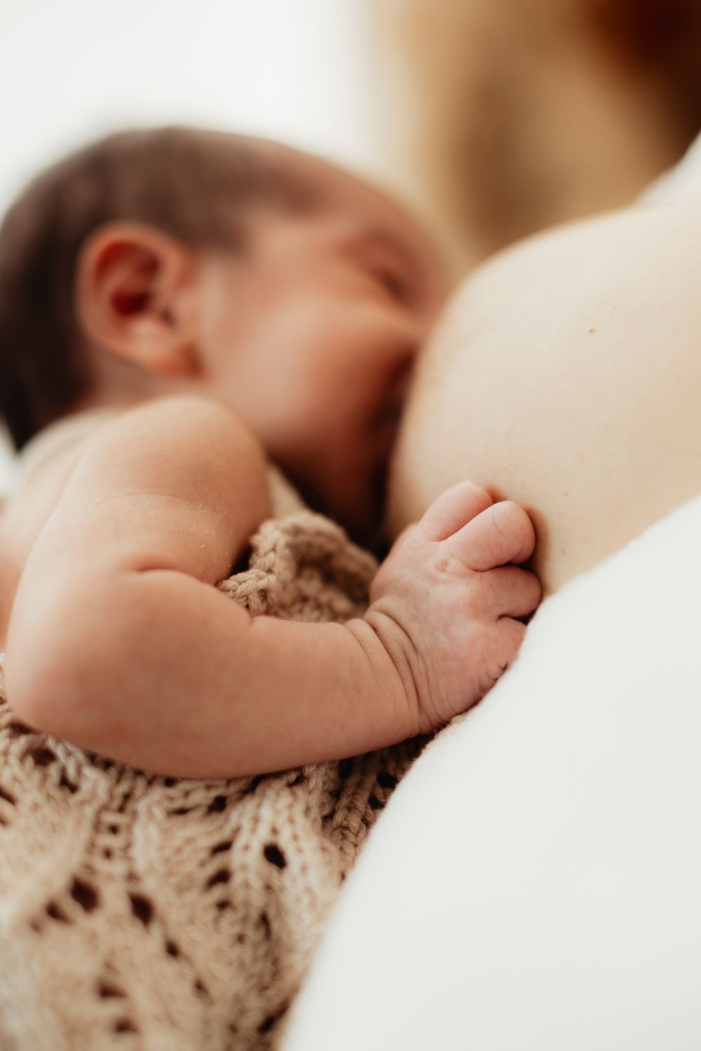 Newborn baby breastfeeding, tiny hand resting on mother's breast, wrapped in a soft knit blanket.