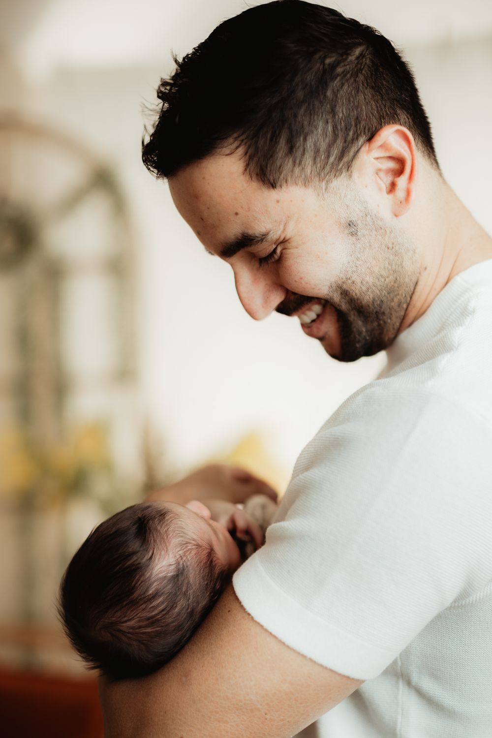 A smiling young father with short dark hair and stubble joyfully holds his newborn baby in a bright home setting.