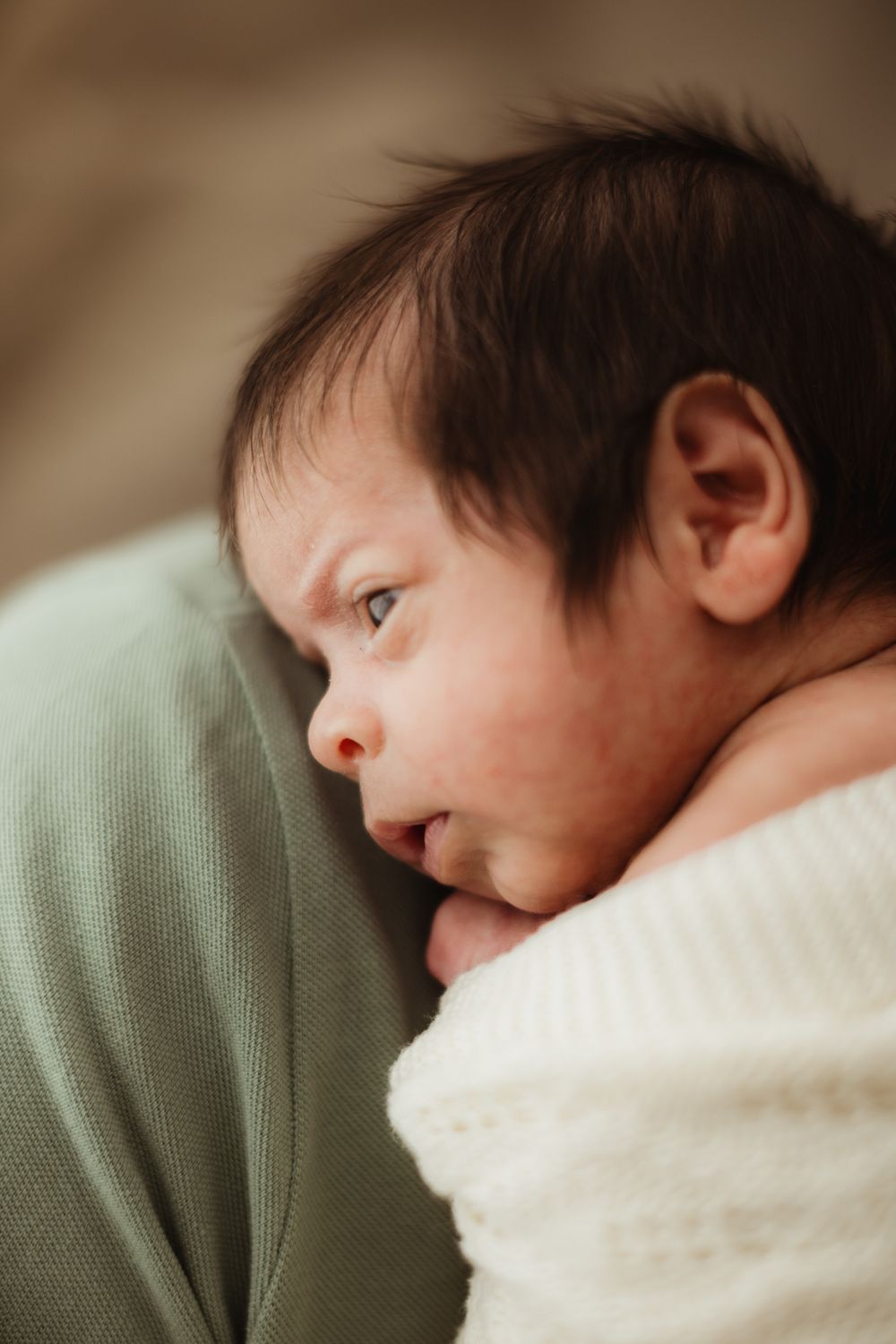 A newborn baby with dark hair wrapped in a white knit blanket resting against a green cloth.