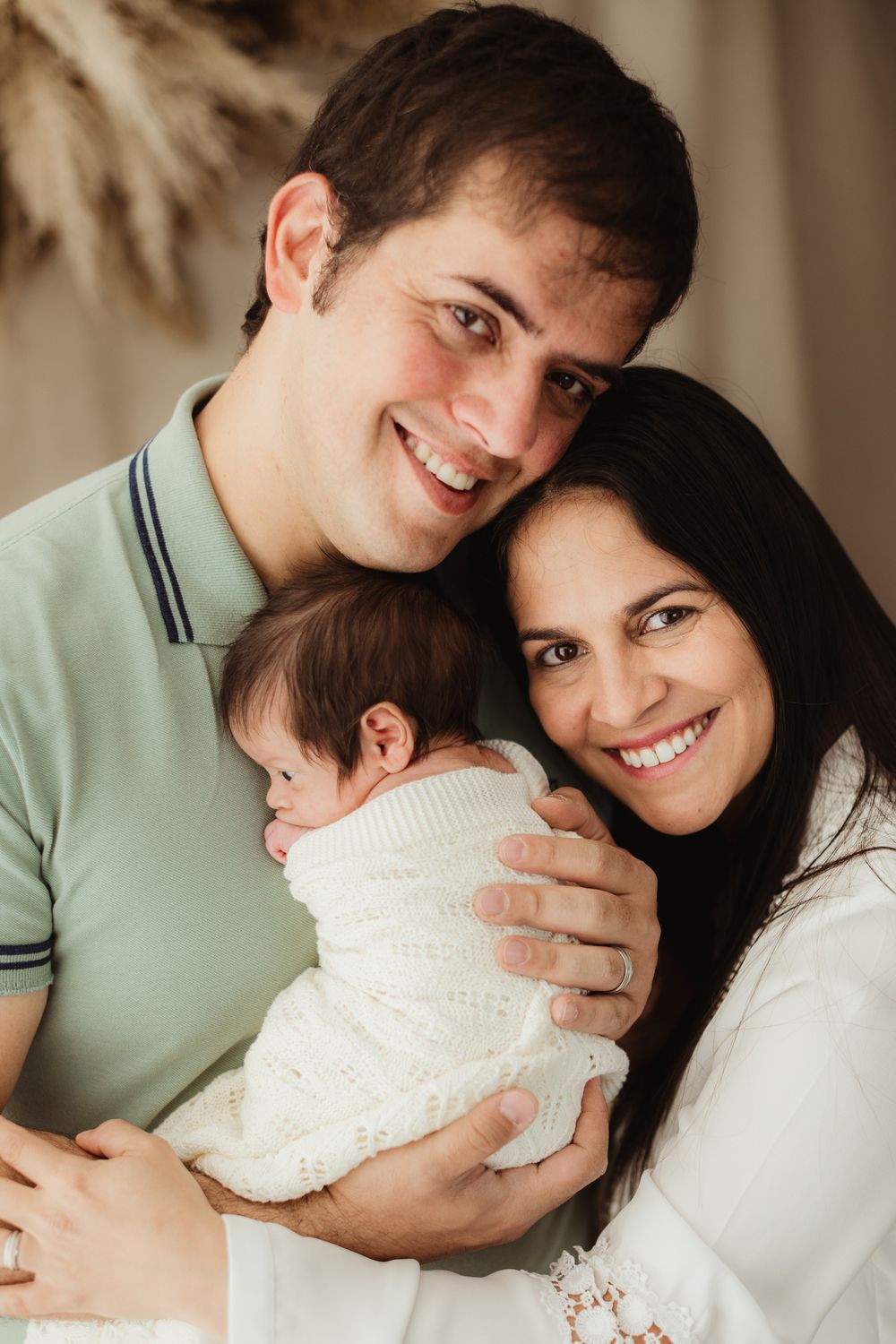 Happy couple smiling and holding their newborn baby wrapped in a white knit blanket in a warm studio setting.