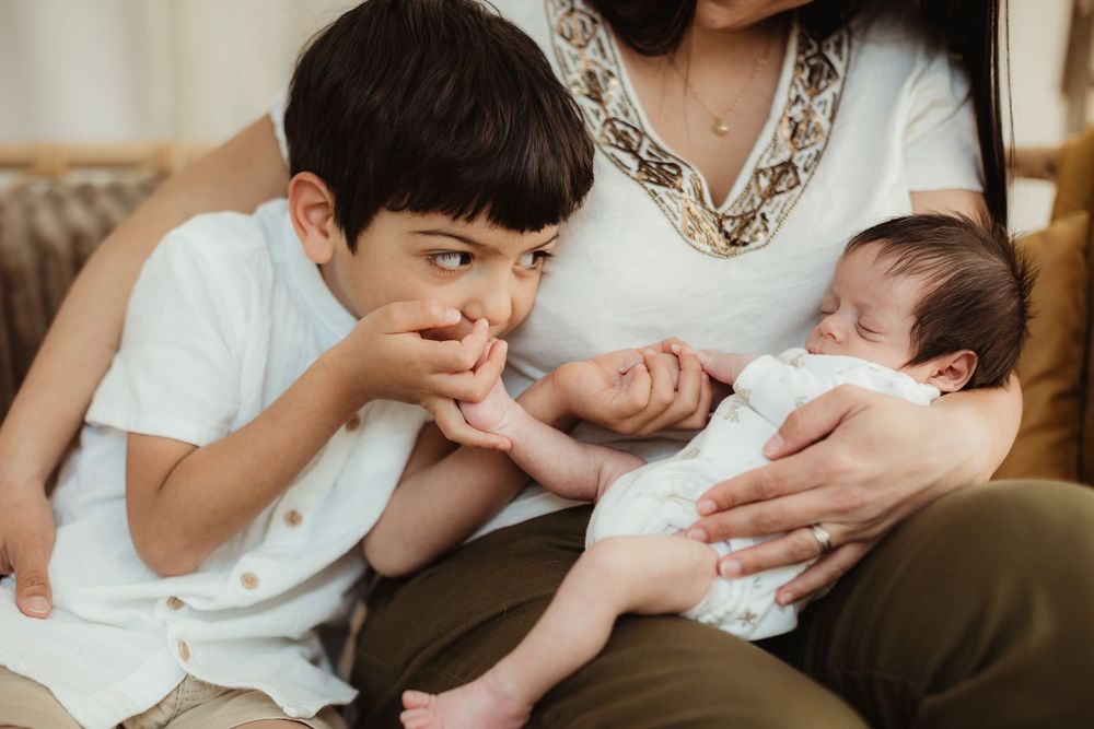 A young boy gazes curiously at a sleeping newborn baby held by their mother on a couch.