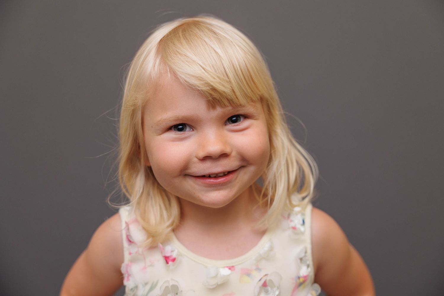 A portrait of a young child with blonde hair smiling against a grey background.