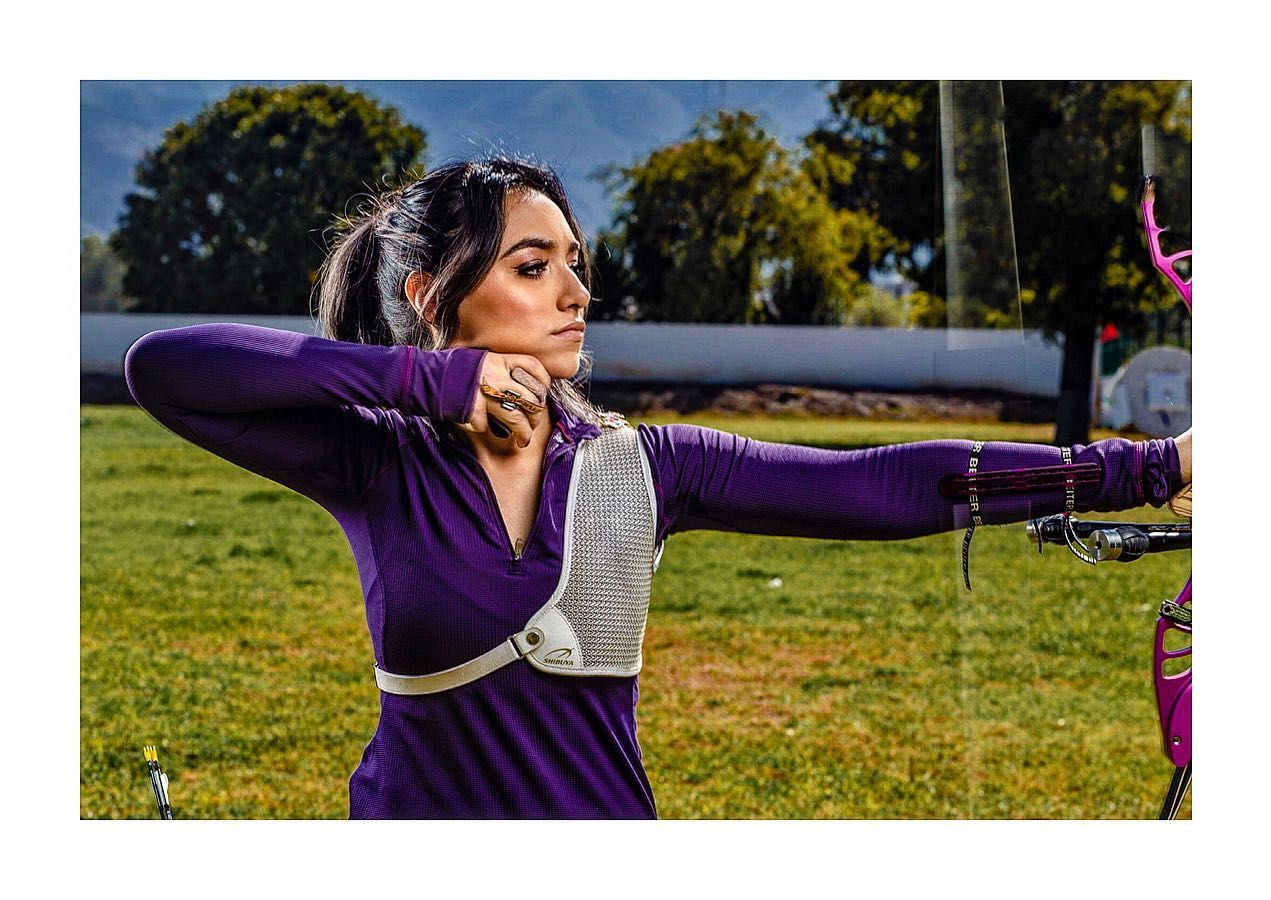 Woman in purple attire practicing archery on a sunny grass field.
