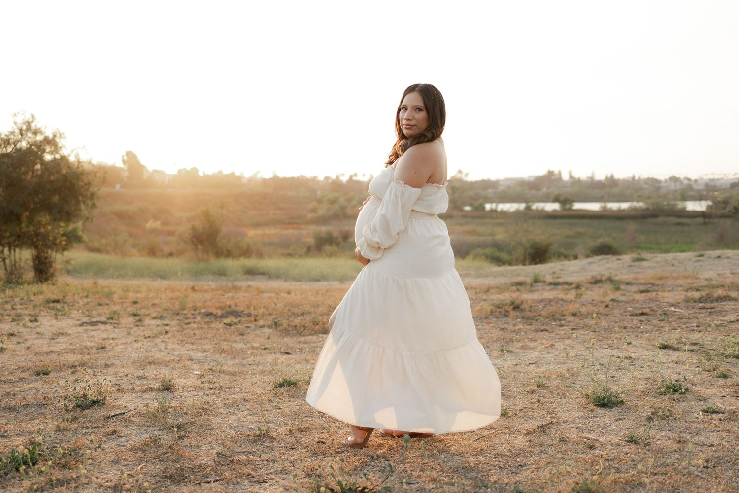 Pregnant woman in a white dress standing outdoors at sunset.