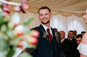 Wedding guest in dark suit with red tie smiles during indoor reception with decorative lighting and floral decor.