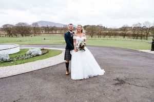 Newlyweds pose together on path with scenic mountain landscape and winter trees in background.