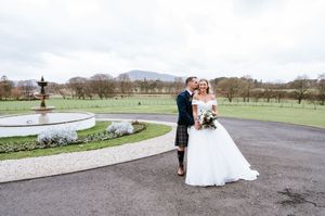 A couple in traditional wedding attire pose together on a curved driveway with mountains visible in the background.