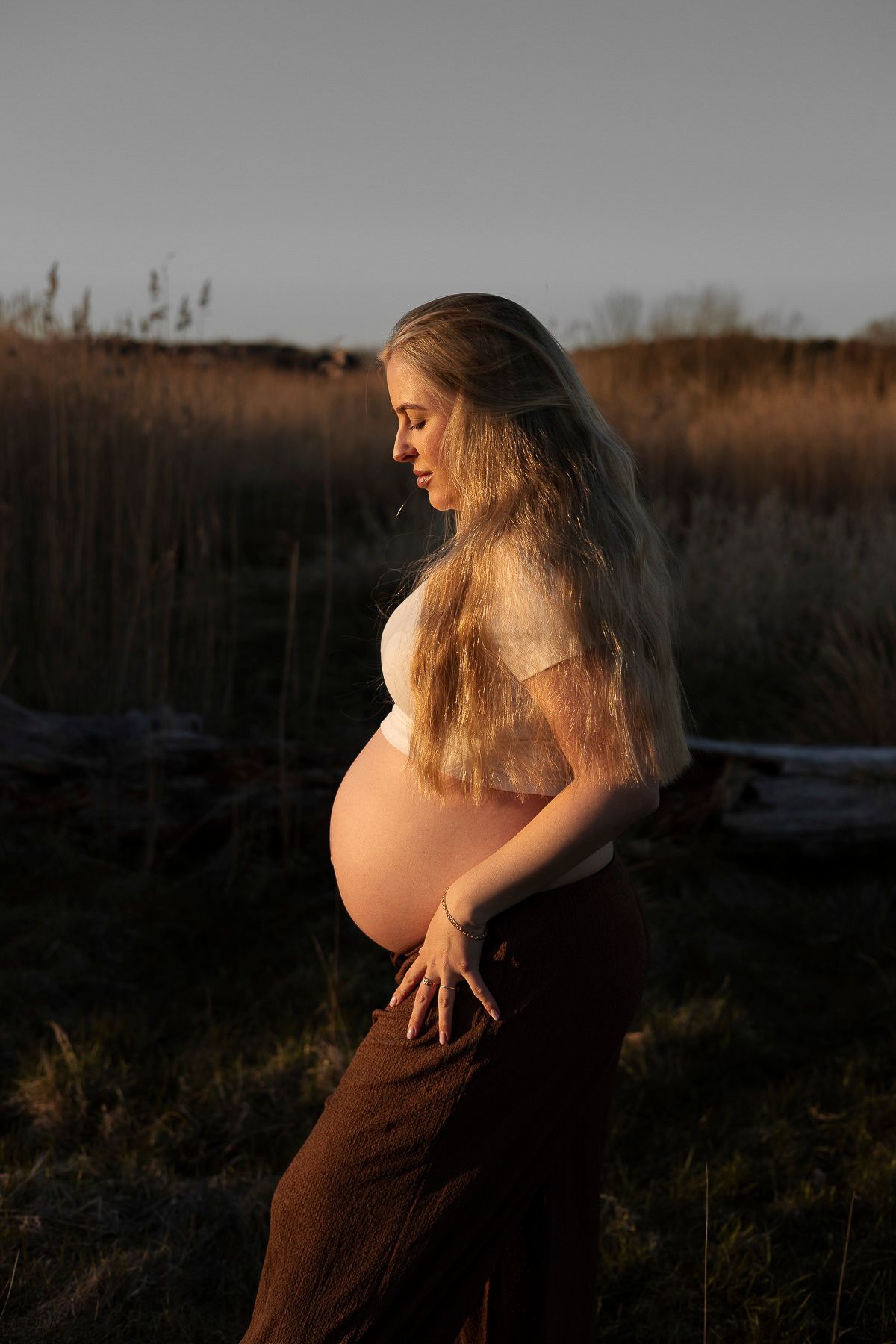 bild tagen från sidan av en gravid kvinna. på sig har hon en vit t-shirt och bruna byxor. solens gyllene sken lyser på henne.