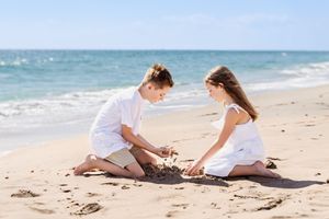 Kids in white clothing examining seashells and playing in the sand at the beach.