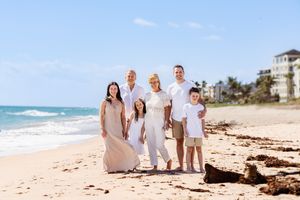 Family dressed in white clothing poses together on a sunny beach with ocean waves in background.