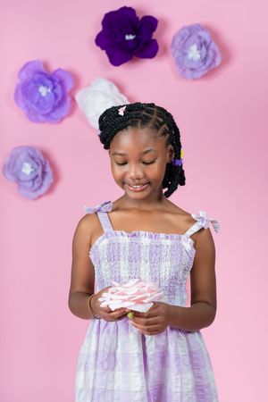 Young girl in white smocked dress poses with cupcake against pink background with purple flowers.