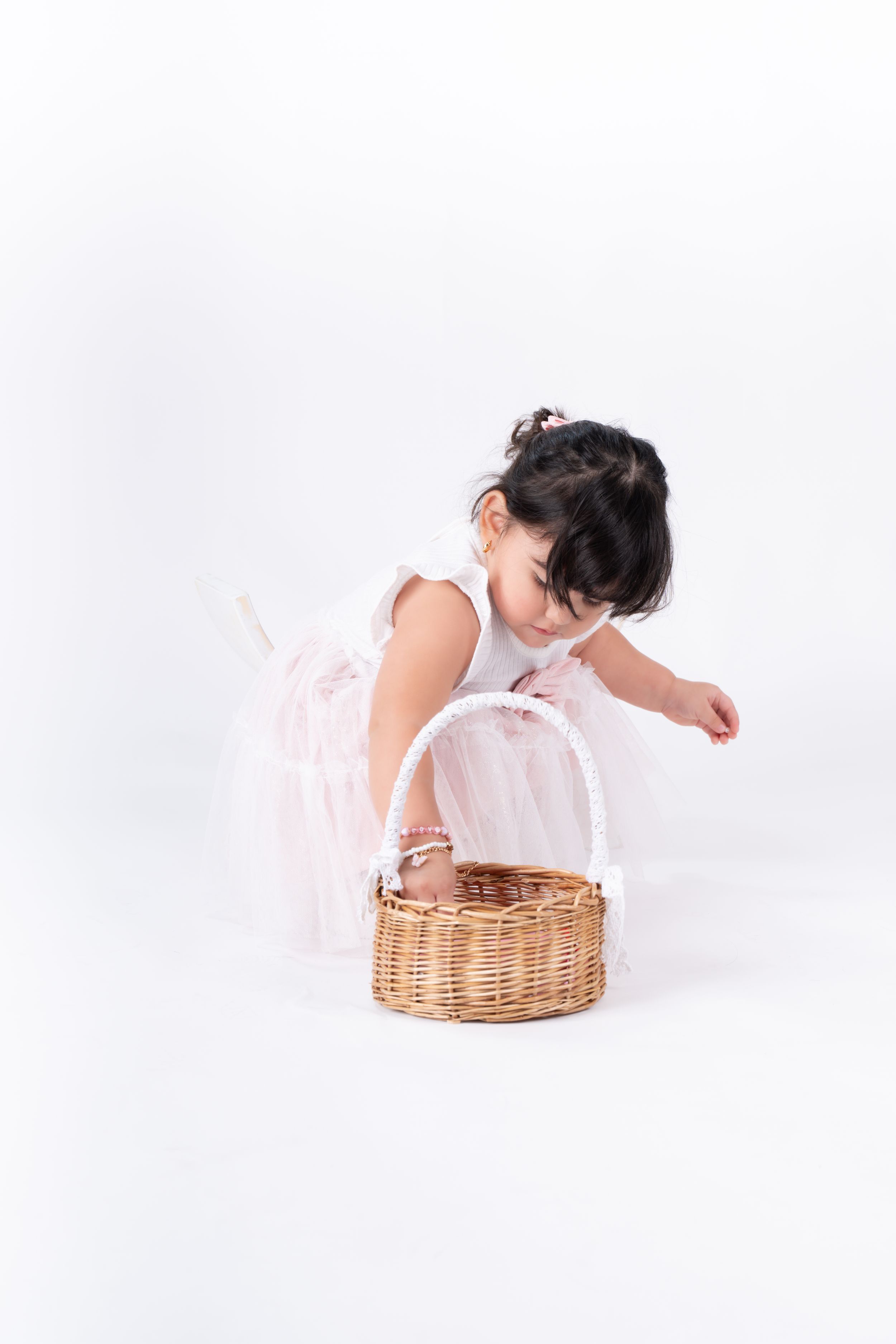 Child in white dress lying near wicker basket on white background.