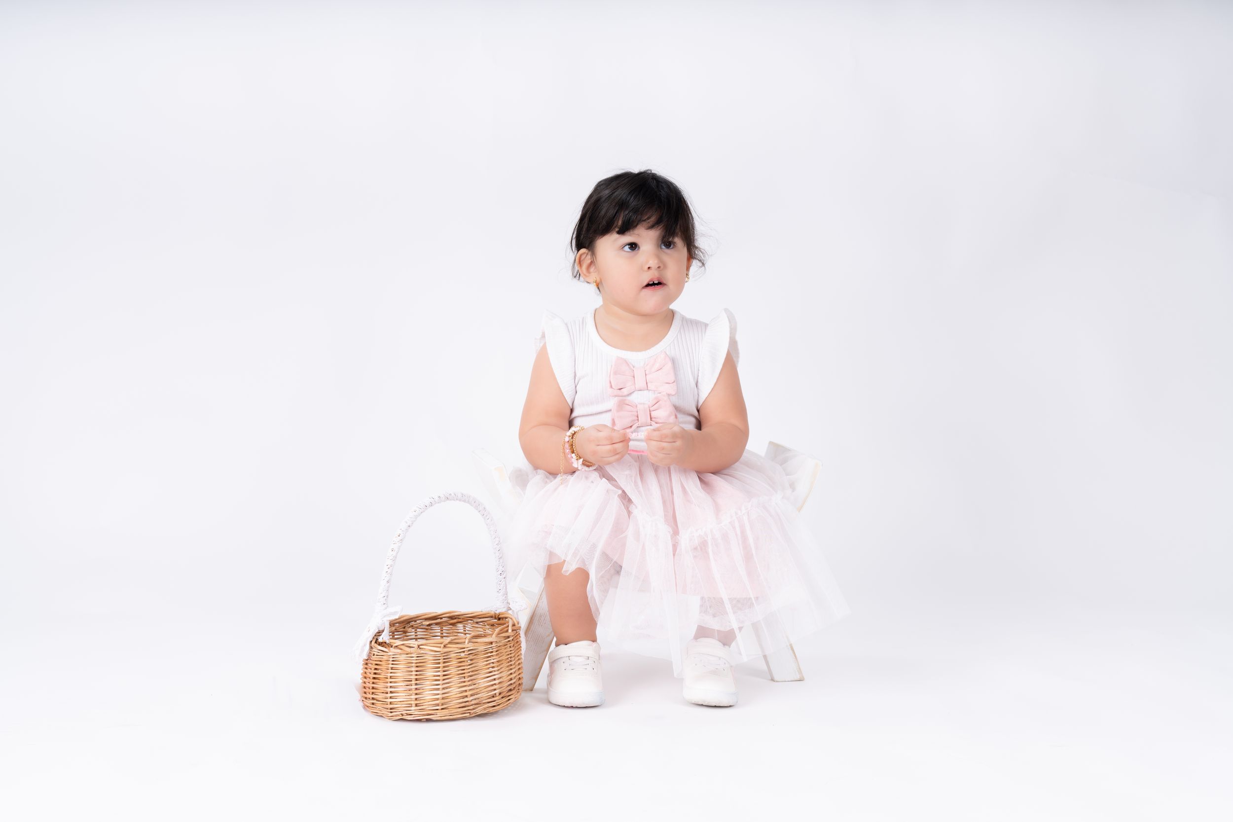 Child in white dress posing with wicker basket against white backdrop.