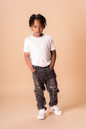 Child in white t-shirt and distressed jeans posing against beige backdrop.